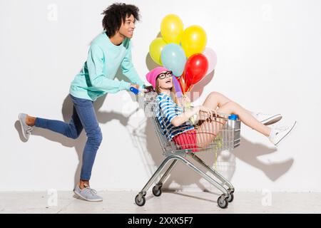 Passer du bon temps ensemble. Heureux jeune homme portant sa belle petite amie dans le panier et souriant tout en courant sur fond gris Banque D'Images
