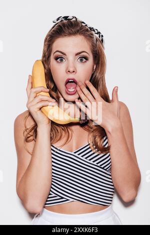 Commérages fruités. Belle jeune femme tenant une banane comme un téléphone et couvrant sa bouche avec la main tout en se tenant debout sur fond blanc Banque D'Images