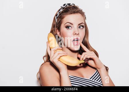 Qu'avez-vous dit ? Belle jeune femme tient la banane comme un téléphone tout en se tenant debout sur fond blanc Banque D'Images