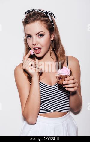 Voler un peu de douceur. Belle jeune femme dégustant un cupcake et regardant la caméra tout en se tenant debout sur fond blanc Banque D'Images