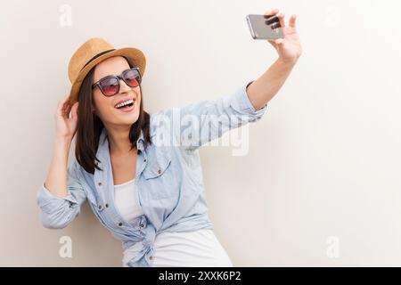 Collecte de souvenirs. Portrait de belle jeune femme dans des lunettes ajustant son chapeau tout en faisant selfie et debout sur fond brun Banque D'Images