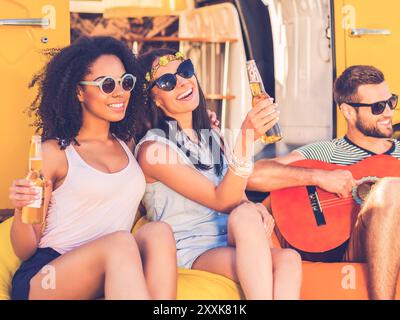 Temps sans soucis avec des amis. Deux jeunes femmes joyeuses tenant des bouteilles avec de la bière et souriant tandis que l'homme joue de la guitare avec minifourgonnette en arrière-plan Banque D'Images