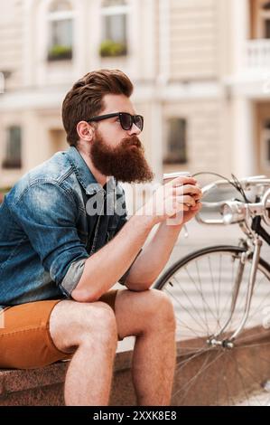 Rester calme. Réfléchi jeune homme barbu tenant une tasse de café tout en étant assis près de son vélo à l'extérieur Banque D'Images