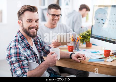 Faire le travail. Beau jeune homme tenant une tasse de café et souriant tout en étant assis au bureau dans le bureau avec ses collègues travaillant dans le BA Banque D'Images