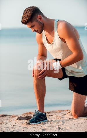 Douleur au genou après la course. Jeune homme musclé confiant massant le genou avant de courir debout au bord de la mer Banque D'Images