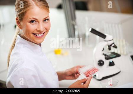 Confiant dans ses recherches. Vue de dessus de la femme scientifique heureuse en uniforme blanc tenant la plaque de microtitration et regardant la caméra tout en étant assis à son w Banque D'Images