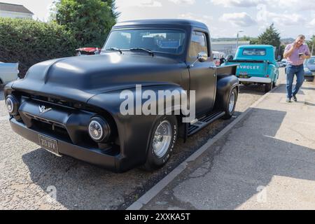Rayleigh, Royaume-Uni. 25 août 2024. Un pick-up Ford noir mat des années 1950 et un camion Chevrolet turquoise sont garés en bord de route lors d'un rassemblement de véhicules classiques. La scène capture le style rétro, les contrastes de restauration et l'intérêt du public pour le patrimoine automobile américain. Un piéton marche à proximité, ajoutant un sentiment de vie urbaine décontractée. Voiture américaine et camion se rencontrent au Farm Fresh garage. L'événement permet aux propriétaires d'apporter leurs véhicules pour que les visiteurs puissent regarder de près. Penelope Barritt/Alamy Live News Banque D'Images