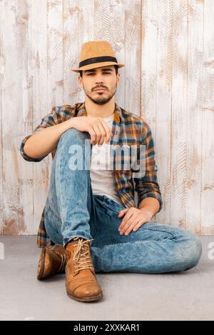 Calme et confiant. Beau jeune homme portant un chapeau et regardant la caméra tout en étant assis contre le mur de bois Banque D'Images