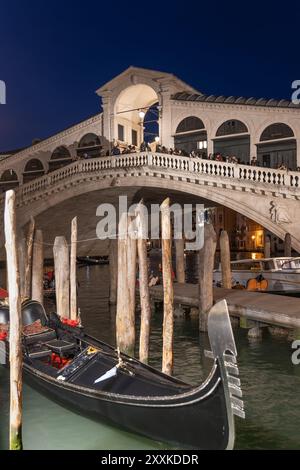 Le pont du Rialto la nuit dans la ville de Venise, Italie. Passerelle en arc de pierre datant de 1591 à travers le Grand canal et gondoles amarrées à un quai. Banque D'Images