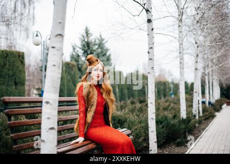 jeune fille avec cheveux rouges dans une robe rouge vif sur un banc dans un parc vide parmi les birches Banque D'Images