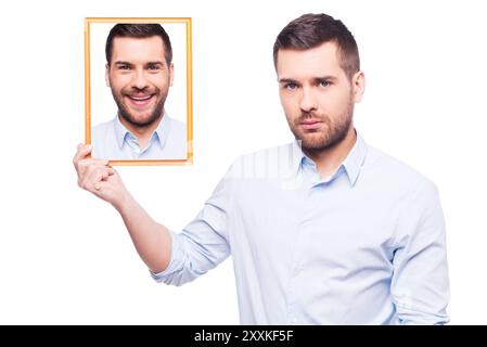 Je souriais... Beau jeune homme en chemise tenant une photo de lui-même avec le visage souriant et l'air triste tout en se tenant debout sur fond blanc Banque D'Images