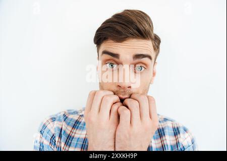 Se sentir si effrayé. Jeune homme nerveux mordant les ongles et regardant la caméra tout en se tenant debout sur fond blanc Banque D'Images