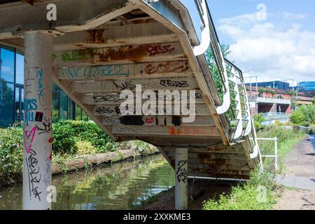 Manchester, Greater Manchester, Royaume-Uni. 24 août 2024 : un pont piétonnier métallique fortement recouvert de graffitis colorés s'étend sur un canal. Les s urbains Banque D'Images