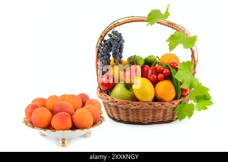 Abricots mûrs dans une assiette en porcelaine et un ensemble de légumes et de fruits dans un panier en osier isolé sur un fond blanc. Collage. Banque D'Images