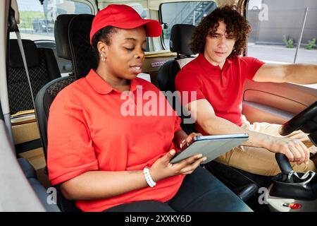 Les livreurs discutent des détails à l'aide d'une tablette lorsqu'ils sont assis dans le véhicule de livraison. Femme portant une casquette rouge et homme aux cheveux bouclés engagés dans la conversation sur leur prochaine livraison Banque D'Images