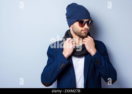 Dans son propre style. Beau jeune homme élégant dans des lunettes de soleil et chapeau ajustant son écharpe et regardant loin tout en se tenant debout sur fond gris Banque D'Images