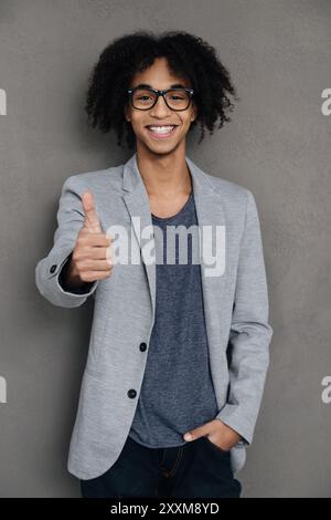 Bravo ! Jeune homme africain joyeux montrant son pouce vers le haut et souriant tout en se tenant debout sur fond gris Banque D'Images