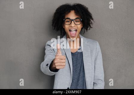 Bravo ! Joyeux jeune homme africain montrant son pouce vers le haut et faisant un visage tout en se tenant debout sur fond gris Banque D'Images