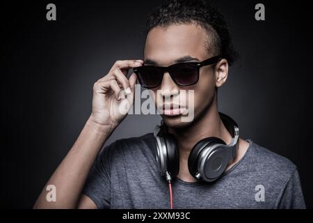 C'est mon style. Portrait d'un jeune homme africain ajustant ses lunettes de soleil et portant des écouteurs tout en se tenant debout sur fond noir Banque D'Images