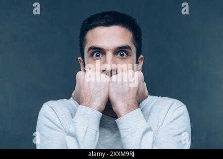 Se sentir choqué. Jeune homme terrifié regardant la caméra et mordant les ongles tout en se tenant debout sur fond gris Banque D'Images