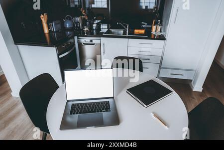 Une configuration de cuisine moderne avec une table ronde blanche, ordinateur portable ouvert avec maquette d'écran, ordinateur portable et stylo. L'arrière-plan présente des appareils de cuisine élégants a Banque D'Images