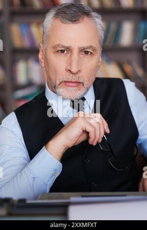 Auteur confiant. Homme senior de cheveux gris confiant en vêtements de forme assis à la machine à écrire et regardant la caméra avec étagère à l'arrière-plan Banque D'Images