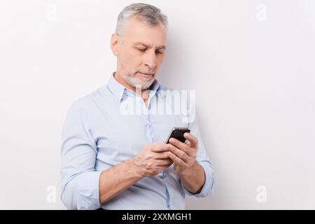 Saisie d'un message professionnel. Homme senior confiant en vêtements de forme tenant le téléphone portable et le regardant tout en se tenant debout sur fond blanc Banque D'Images