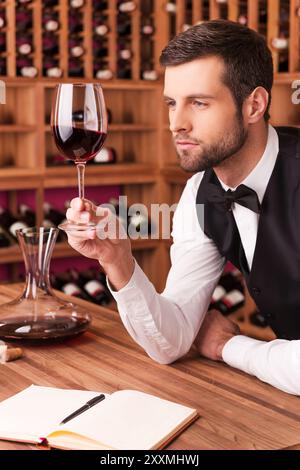 Ce vin est parfait. Sommelier masculin confiant examinant le vin tout en regardant le verre à vin et en se penchant sur la table en bois avec étagère à vin dans le BA Banque D'Images