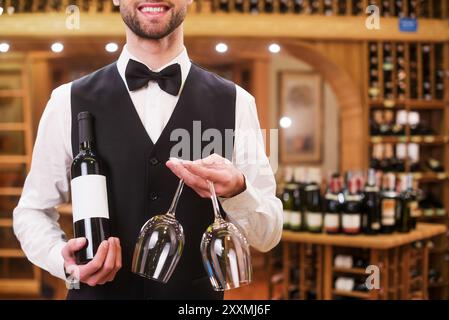 Serveur confiant. Beau jeune homme en gilet et noeud papillon tenant bouteille et verres tout en se tenant debout dans le magasin d'alcool Banque D'Images