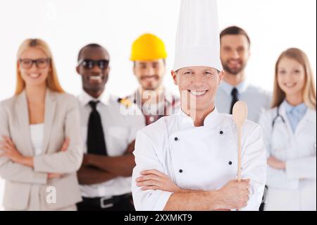 Quand je serai grand, je serai chef. Chef masculin confiant en uniforme gardant les bras croisés et souriant tout en groupe de personnes dans différentes professions standi Banque D'Images