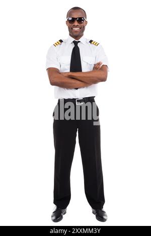 Pilote confiant. Pleine longueur de pilote masculin confiant en uniforme gardant les bras croisés et souriant tout en se tenant debout sur fond blanc Banque D'Images