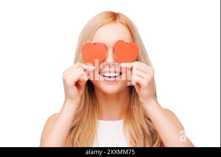 L'amour est un grand sentiment! Belle jeune femme aux cheveux blonds tenant des cartes de saint-valentin en forme de coeur devant ses yeux et souriant tout en se tenant isolé Banque D'Images