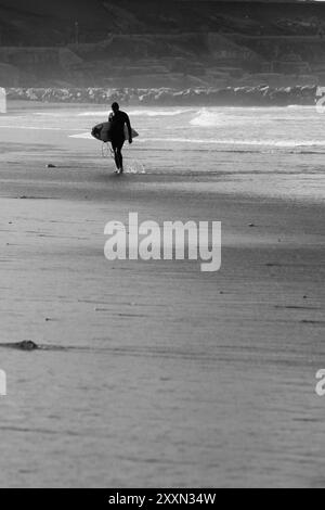 une photo en niveaux de gris d'un surfeur marchant au bord de l'océan à distance Banque D'Images