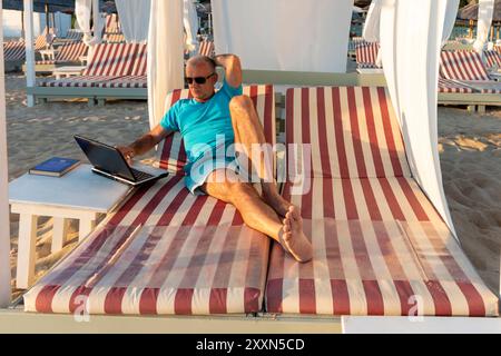 Homme d'affaires d'âge moyen utilisant un ordinateur portable tout en s'allongeant sur le lit de repos sur la plage. Banque D'Images