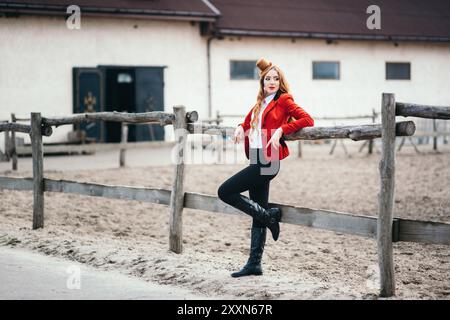 une jeune fille jockey aux cheveux rouges dans un gilet rouge et des bottes hautes noires se tarèrent à la clôture Banque D'Images