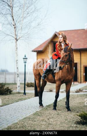 une jeune fille jockey aux cheveux rouges dans un gilet rouge et des bottes noires hautes avec un cheval pour une promenade Banque D'Images