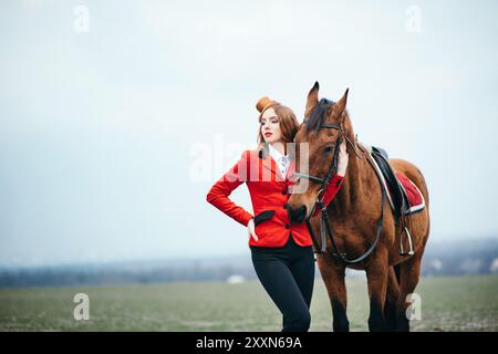 une jeune fille jockey aux cheveux rouges dans un gilet rouge et des bottes noires hautes avec un cheval pour une promenade Banque D'Images