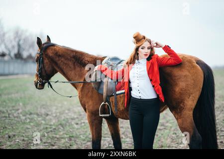 une jeune fille jockey aux cheveux rouges dans un gilet rouge et des bottes noires hautes avec un cheval pour une promenade Banque D'Images