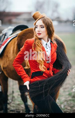 une jeune fille jockey aux cheveux rouges dans un gilet rouge et des bottes noires hautes avec un cheval pour une promenade Banque D'Images