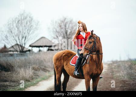 une jeune fille jockey aux cheveux rouges dans un gilet rouge et des bottes noires hautes avec un cheval pour une promenade Banque D'Images