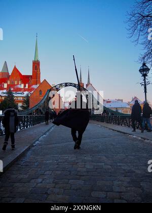 Wroclaw, Pologne - 20 janvier 2024 : une personne vêtue de vêtements sombres traverse un pont pavé avec vue sur les cathédrales et le bâtiment historiques Banque D'Images