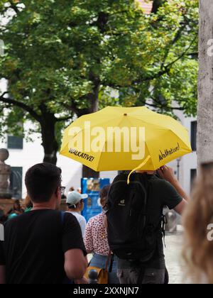 Munich, Allemagne - 3 août 2024 : un groupe de personnes se promenant sous un parapluie jaune vif avec Walkative imprimé dessus, créant un contraste contre Banque D'Images