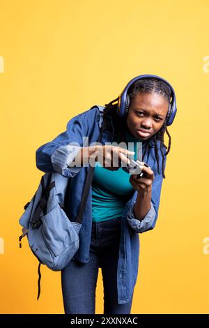 Joueur afro-américain tenant le contrôleur, plissant les yeux, essayant de terminer le niveau du jeu vidéo, portant des écouteurs. Femme jouant avec gamepad, vaincre d'autres joueurs, toile de fond de studio Banque D'Images