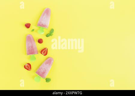 Flat Lay avec une crème glacée au sorbet congelé rose fruité sur un bâton en plastique vert collection avec des coupes de fraise, des feuilles de menthe sur fond jaune. Gelé Banque D'Images