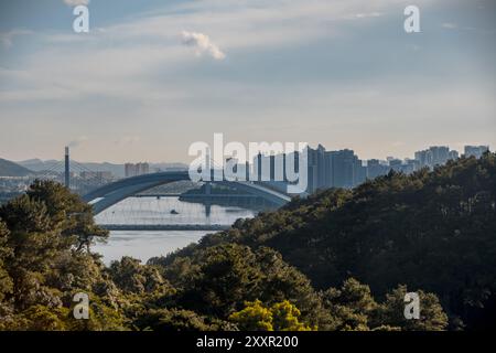 Horizon urbain avec pont moderne sur une rivière, entouré d'arbres verdoyants et sous un ciel partiellement nuageux pendant la journée Banque D'Images