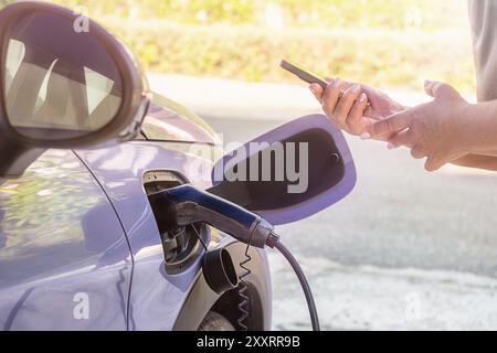 Recharge de voiture électrique. Mains féminines tenant un téléphone près de la voiture Banque D'Images