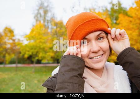 Femme souriante ajuste son bonnet orange tout en profitant d'une belle journée d'automne dans un parc Banque D'Images