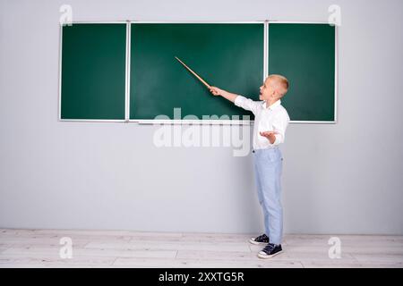 Photo de charmant apprenant gentil garçon intelligent debout dans la salle de classe pointant la réponse sur l'espace vide du tableau vert Banque D'Images