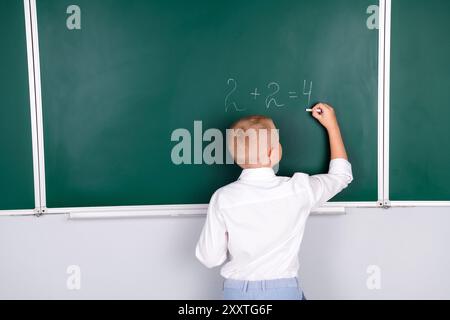 Photo de garçon diligent intelligent dans la salle de classe résoudre la tâche mathématique d'écriture isolé sur fond de tableau vert Banque D'Images