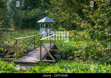 Un petit gazebo dans un parc avec des arbres. Un pont en bois sur la rivière mène à un belvédère. Banque D'Images